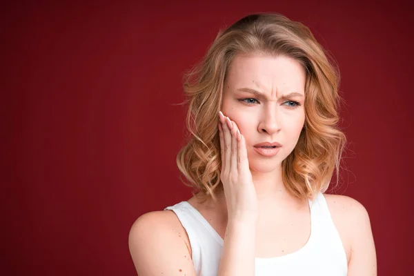 Wanita pirang dengan rambut keriting terlihat bingung atau memiliki sakit gigi di atas tangki putih pada latar belakang merah. — Stok Foto