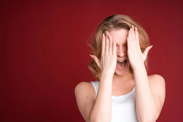 Blond žena křičí a zakrývá si oči s oběma rukama v bílém tílku na červeném pozadí. Vyděšený. — Stock fotografie