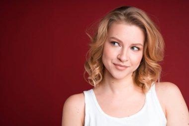 Güzel sarışın kadın kırmızı arka planda beyaz kolsuz bluzuyla yan yana bakıyor..