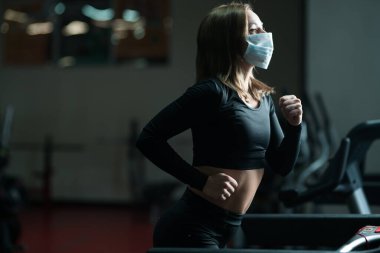 Koronavirüs salgını sırasında spor salonunda koşu yaparken koruyucu yüz maskesi takan bayan sporcu. Koşu bandında ısınma turu.