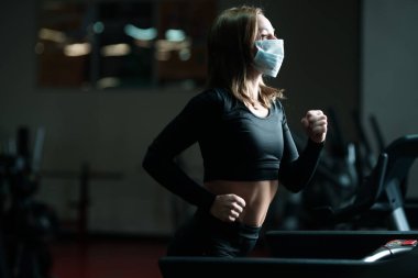 Koronavirüs salgını sırasında spor salonunda koşu yaparken koruyucu yüz maskesi takan bayan sporcu. Koşu bandında ısınma turu.