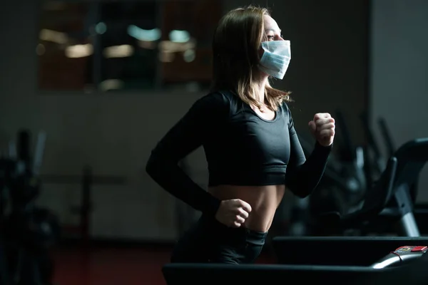 Atleta femenina corriendo en pista mientras usa mascarilla protectora en un gimnasio durante la epidemia de coronavirus. calentamiento en una cinta de correr —  Fotos de Stock