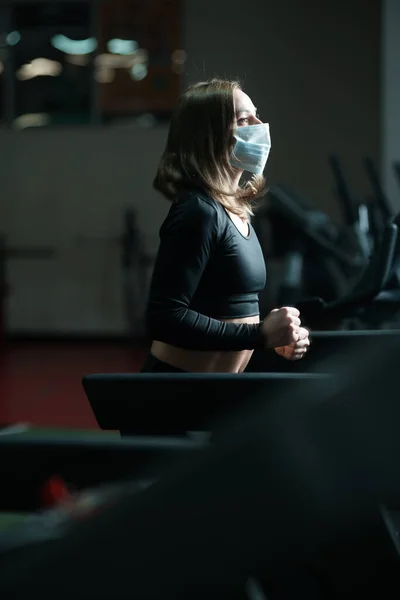 Una chica entrenadora de fitness en una máscara médica se dedica a una cinta de correr, desarrolla los pulmones durante una pandemia de coronavirus. —  Fotos de Stock