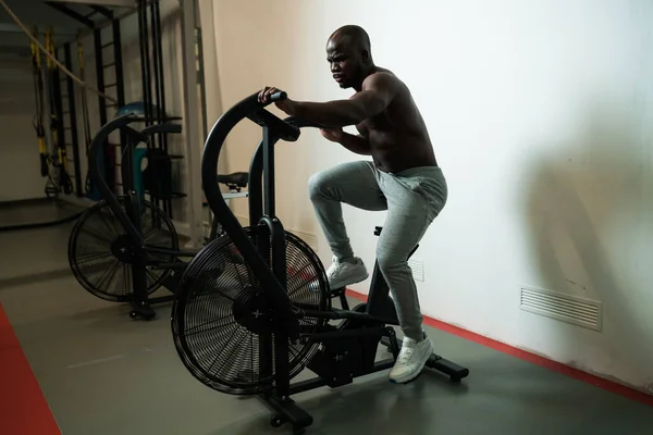 Νέοι ισχυρή εφαρμογή μυϊκή Αφροαμερικανός άνθρωπος με μεγάλους μυς κάνει άσκηση καρδιο στο εσωτερικό ποδήλατο αέρα στο γυμναστήριο ως σκληρή προπόνηση σταυρό. Έννοια της crossfit, αθλητισμός. — Φωτογραφία Αρχείου