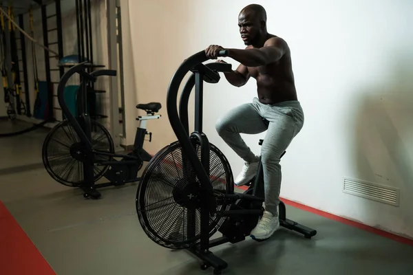 Ισχυρή εφαρμογή μυϊκή αφρικανική Αμερικανός άνθρωπος με μεγάλους μυς κάνει άσκηση καρδιο στο εσωτερικό ποδήλατο αέρα στο γυμναστήριο ως σκληρή προπόνηση σταυρό. Έννοια της crossfit, αθλητισμός — Φωτογραφία Αρχείου