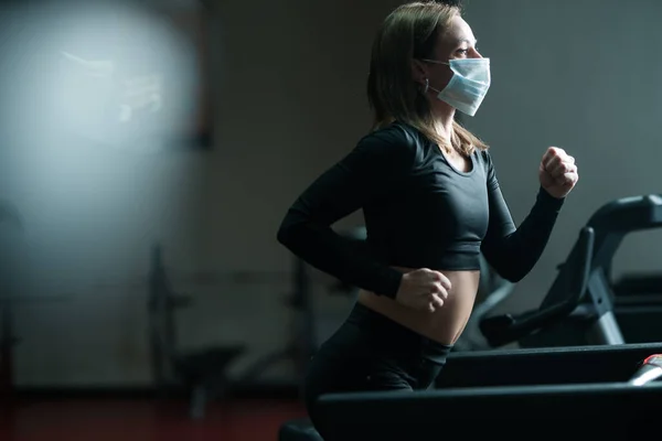 Una chica entrenadora de fitness en una máscara médica se dedica a una cinta de correr, desarrolla los pulmones durante una pandemia de coronavirus —  Fotos de Stock