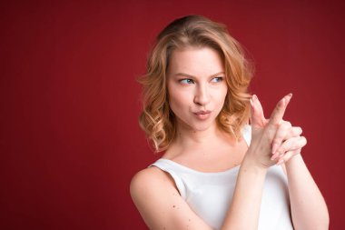Blond woman with curly hair showing finger gun in white tank top on red background.