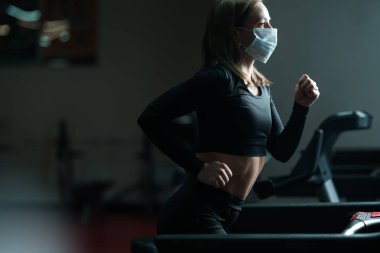Spor kadın spor salonunda koşu bandında antrenman yapıyor ve küresel covid-19 virüsü salgınında kendini koronavirüsten korumak için maske takıyor.
