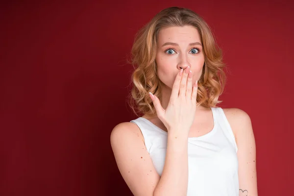 Cute woman with curly blond hair covering mouth with hand in white tank top on red background. — Zdjęcie stockowe