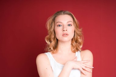 Blond woman with curly hair puts her hand on chest isolated on red background.
