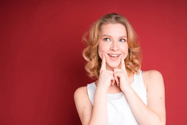 Blondýny žena s kudrnaté vlasy dělat úsměv s prsty v bílé tílko top izolované na červeném pozadí. — Stock fotografie