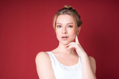 Cute blond woman with bun looks dreamy or thoughtful in white tank top isolated on red background.