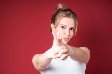 Blond woman with curly hair showing finger gun in white tank top on red background.