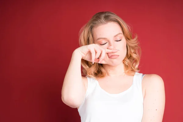 Blond žena s kudrnaté vlasy otírá nos s prstem v bílé tílko top izolované na červeném pozadí. — Stock fotografie