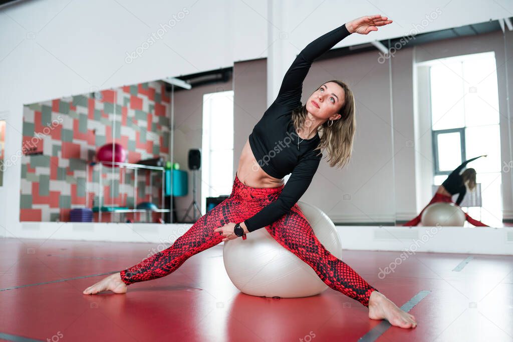 A girl in a black top and red leggings does exercises for stretching the inner thigh on a gymnastic ball in a fitness studio
