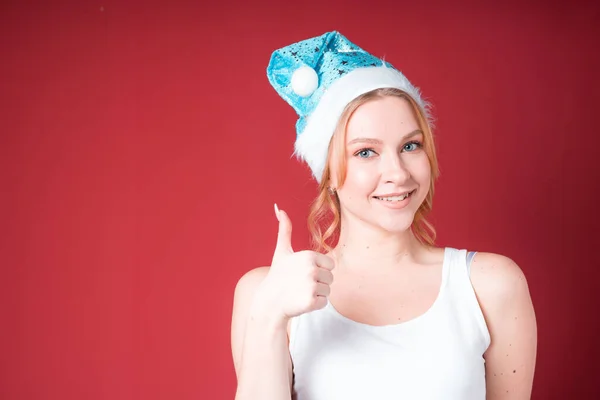 Χαριτωμένο ξανθιά γυναίκα χαμογελά και δείχνει τον αντίχειρα επάνω σε λευκό δεξαμενή κορυφή και μπλε καπέλο Χριστούγεννα απομονώνονται σε κόκκινο φόντο. — Φωτογραφία Αρχείου