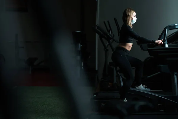 Flickan är ensam i gymmet på ett löpband. Utbildning i medicinsk mask och handskar under coronaviruspandemin — Stockfoto