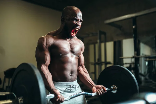 Emocional muscular afro-americano homem tensionando músculos, halterofilista grita e levantando uma barra pesada — Fotografia de Stock