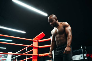 Alt manzara. Seçkin kasları ve çıplak gövdesi olan Afro-Amerikan bir adam spor salonundaki bir boks ringinin arka planında duruyor.