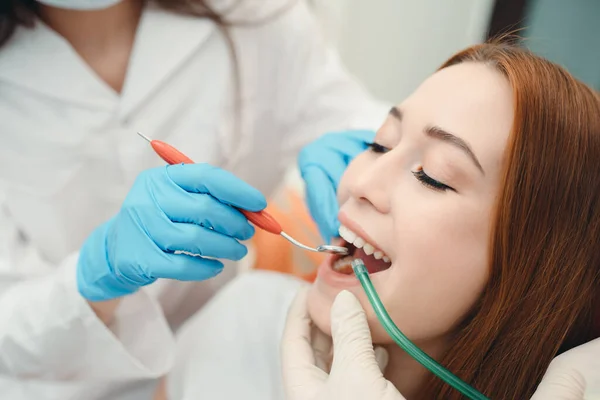 A dentist examines the teeth of a red-haired girl sitting in a chair. Portrait of a girl with saliva ejector at a dentist appointment