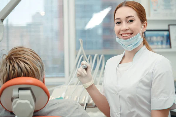 Aantrekkelijke vrouwelijke tandarts in een witte medische jurk met een tandheelkundig instrument in de hand poserend voor de camera naast de patiënt in een stoel — Stockfoto