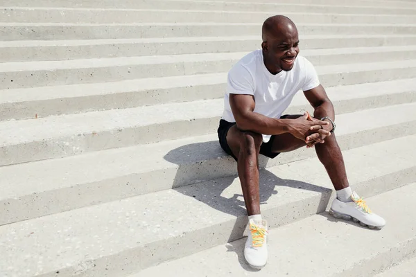 Hombre afroamericano en camiseta blanca y pantalones cortos negros descansando después de un duro día de entrenamiento al aire libre. — Foto de Stock