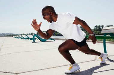 Beyaz tişörtlü ve siyah şortlu siyah adam dışarıda elastik bantla egzersiz yapıyor..