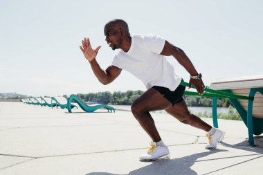 Beyaz tişörtlü ve siyah şortlu siyah bir adam dışarıda lastik bantla egzersiz yapıyor ve çığlık atıyor..