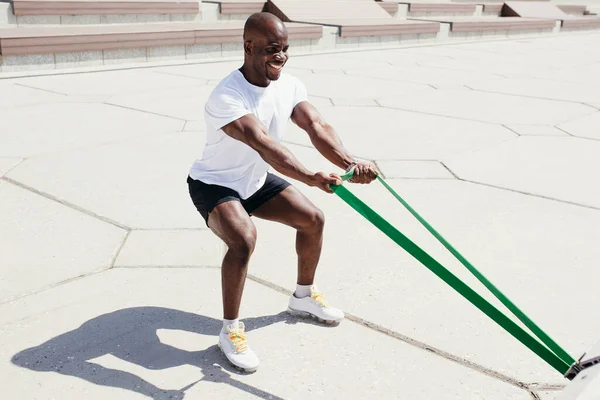 Afro hombre americano en camiseta blanca y pantalones cortos negros haciendo ejercicio en sus manos en cuclillas con cinta elástica de fitness afuera. —  Fotos de Stock