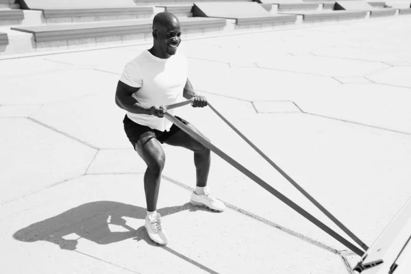 Hombre afroamericano en camiseta blanca y pantalones cortos negros haciendo ejercicio en sus manos ocupándose con cinta elástica de fitness afuera. Monocromo foto. —  Fotos de Stock