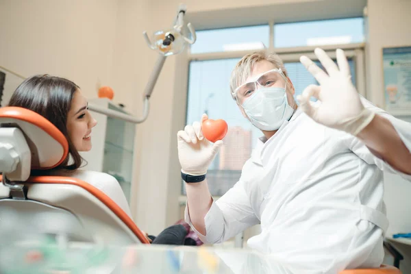 Een tandarts in medische handschoenen houdt een rood speelgoedhart in zijn hand naast een vrouw die op een bank ligt — Stockfoto