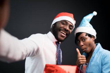 Afrikalı Amerikalılar. Kırmızı Noel Baba şapkalı bir adam ve mavi şapkalı bir kadın hediye kutusuyla selfie çekiyorlar.
