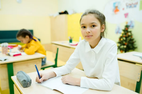 Mała dziewczynka w białej koszuli z zebranymi włosami siedzi przy biurku i pisze w notatniku — Zdjęcie stockowe