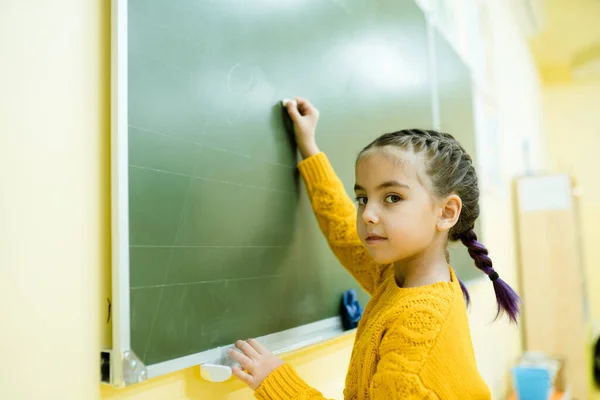Dziewczyna w żółtym swetrze w warkoczu pisze temat na zielonej tablicy. Szkoły — Zdjęcie stockowe