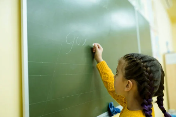 Dziewczyna w żółtym swetrze w warkoczu pisze temat na zielonej tablicy. Szkoły — Zdjęcie stockowe