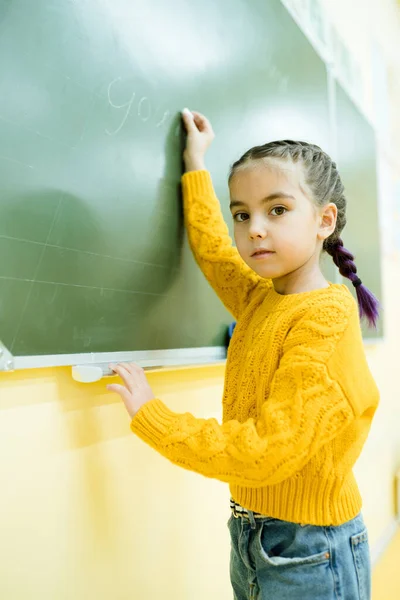 Mały uczeń w żółtym swetrze patrzy w kamerę podczas pisania zadania w lekcji — Zdjęcie stockowe