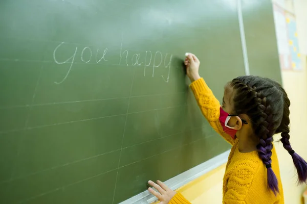 Mały uczeń w żółtym swetrze patrzy w kamerę podczas pisania zadania w lekcji — Zdjęcie stockowe