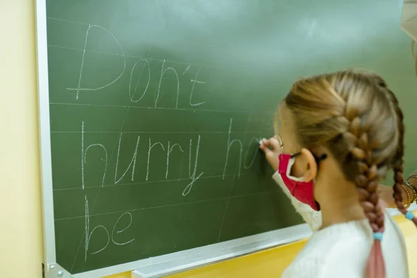 Dziewczyna z warkoczami w masce ochronnej na lekcji pisze kredą na tablicy. — Zdjęcie stockowe