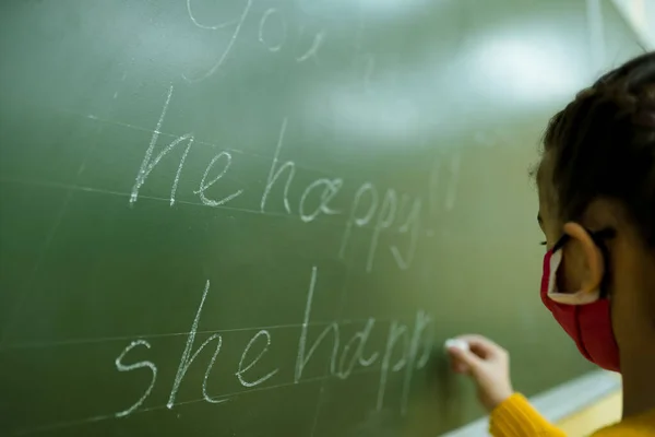 Dziewczyna w masce medycznej pisze na tablicy listy kredą. Lekcje w szkole — Zdjęcie stockowe
