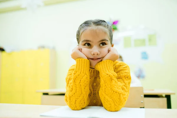 Małe dziecko w żółtym swetrze siedzi przy szkolnym biurku z łokciami na rękach. — Zdjęcie stockowe
