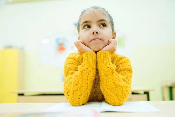 Małe dziecko w żółtym swetrze siedzi przy szkolnym biurku z łokciami na rękach. — Zdjęcie stockowe