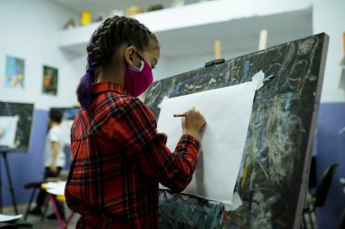 Tıbbi maskeli, ekose gömlekli bir kız kağıt tutacağının yanında duruyor ve elinde bir kalem tutuyor.