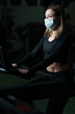 Siyah atletli ve koruyucu maskeli bir kadın spor salonunda otururken spor yapıyor.