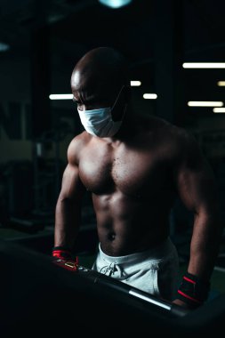 Koruyucu maskeli çıplak gövdeli bir adam koşu bandında egzersiz yaparak atletik fiziği gösteriyor.