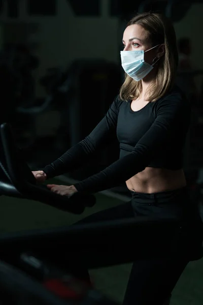 Mujer con camiseta negra y polainas en una máscara protectora realiza un ejercicio mientras está sentada en el gimnasio —  Fotos de Stock