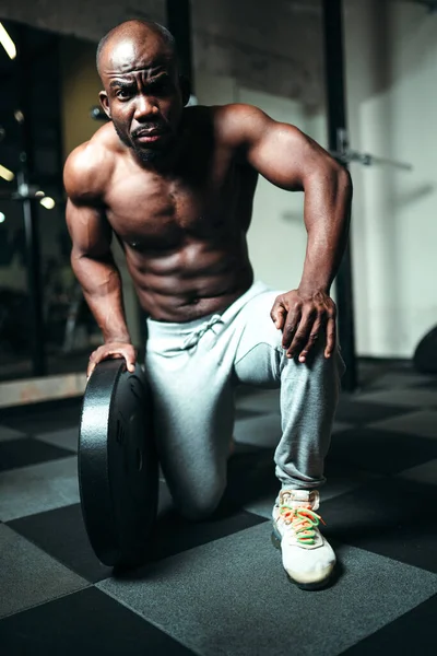 Masculino Africano americano powerlifting treinador sentado em um joelho no chão no ginásio com um disco de barra na mão — Fotografia de Stock