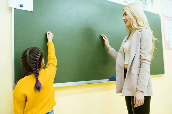 Nauczyciel z długimi włosami stoi przy tablicy z uczniem piszącym kredą — Zdjęcie stockowe