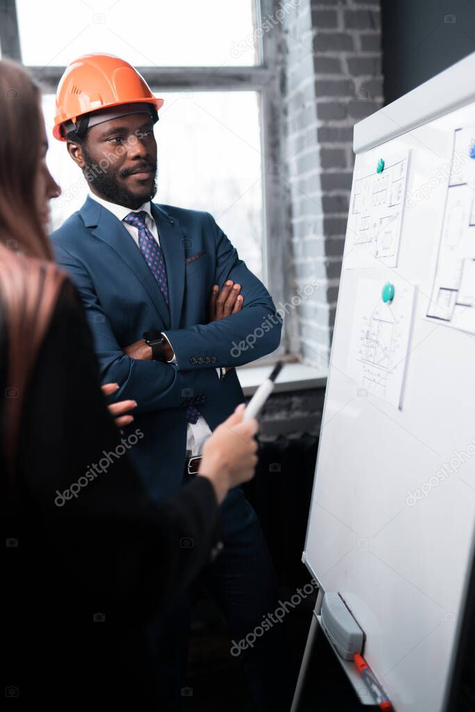 African American man in formal suit stands near a magnetic board and is interested in the plan and diagrams of buildings