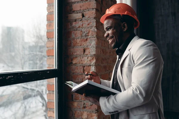 Афро-американский инженер в оранжевом строительном шлеме и деловом костюме держит папку с проектом нового бизнес-центра в руках, стоит на строительной площадке — стоковое фото