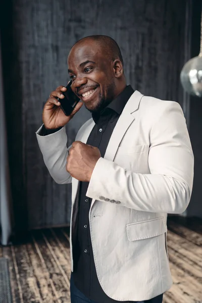 Um homem africano empresário americano com um casaco leve e camisa preta fala ao telefone e se alegra com a conclusão de um acordo — Fotografia de Stock